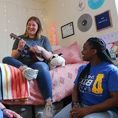 a student plays a ukulele in their room while another student sits on the floor